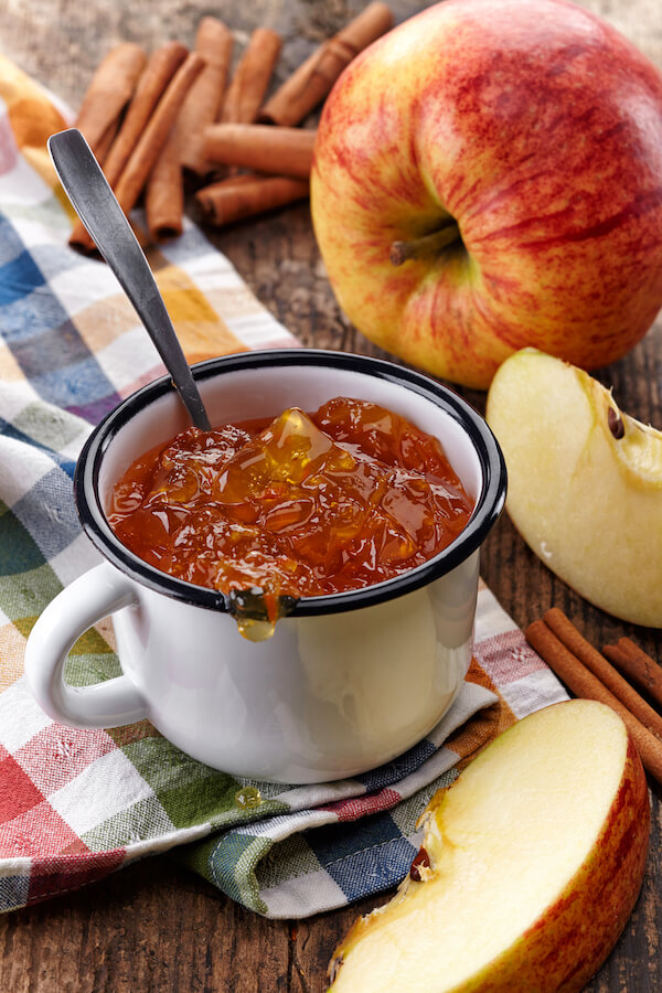 Homemade apple preserves in a white mug on a table with apples and cinnamon sticks.