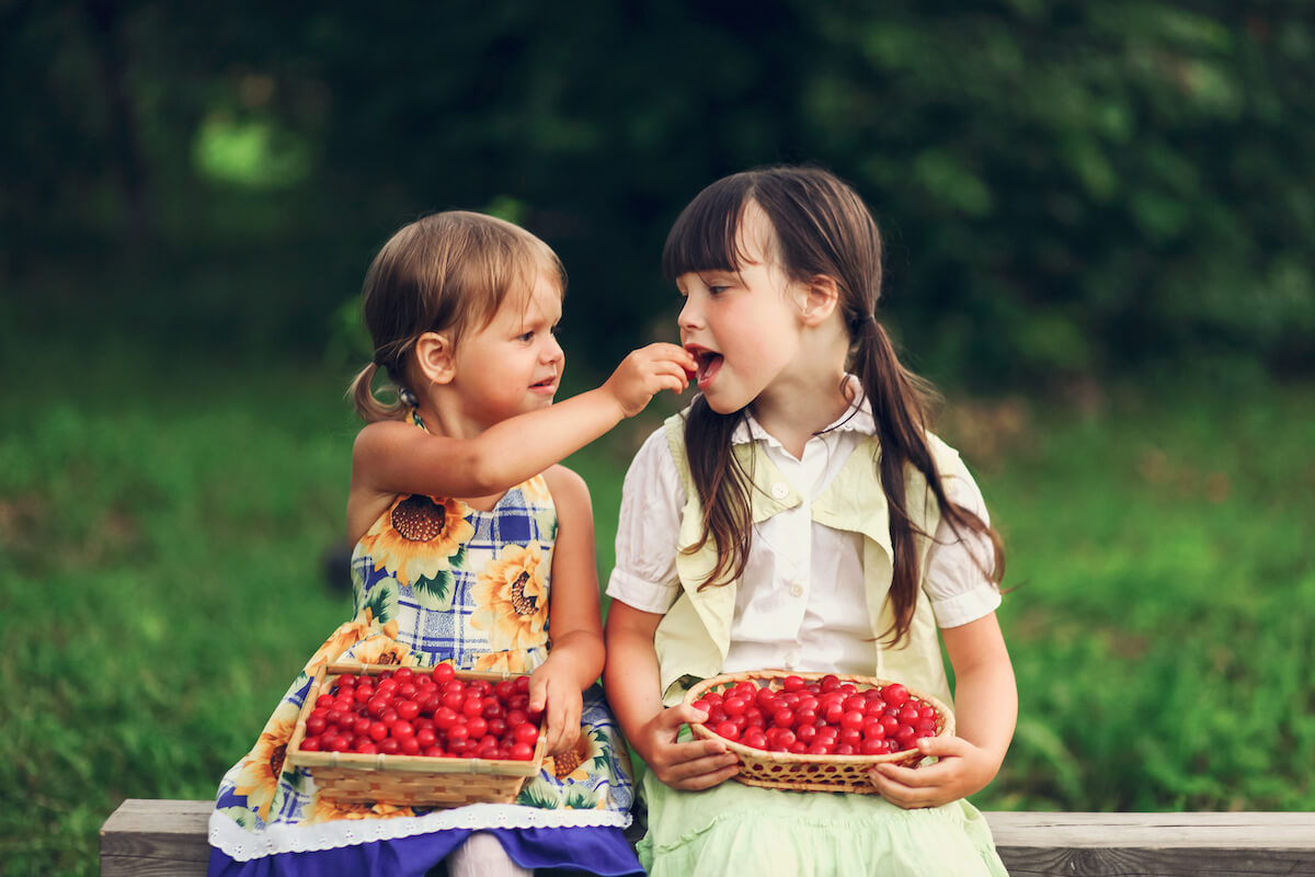 Two girls holding baskets of cherries, one feeding one to the other.
