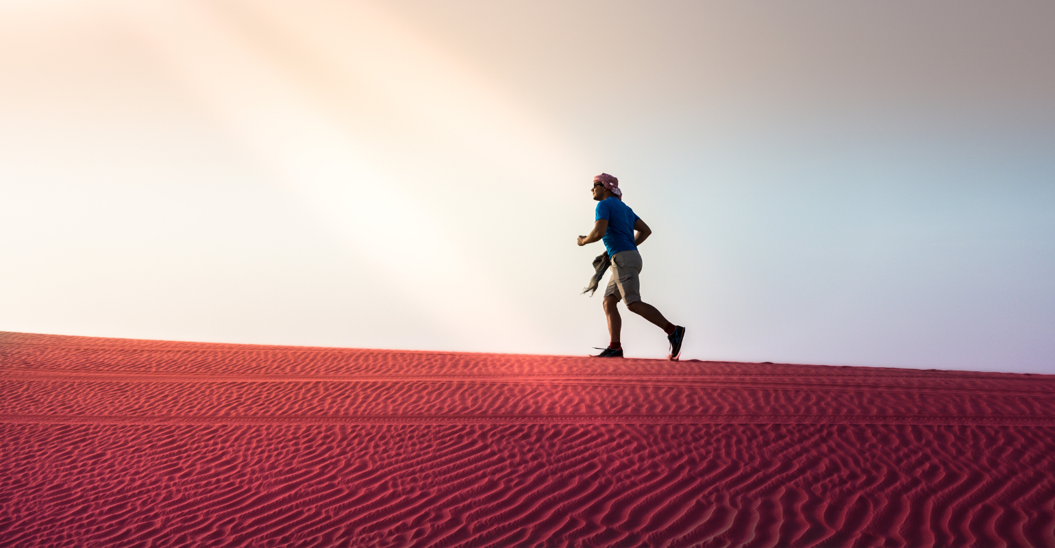 An adventrous man in shorts, t-shirt, and headwrap running across rippling red sand.